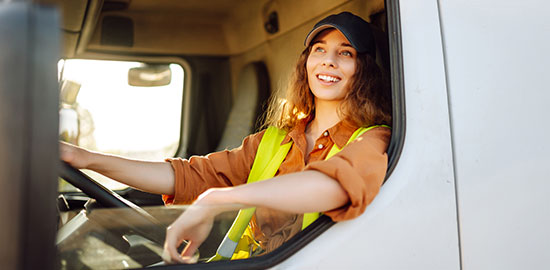 Eine Fahrerin mit Warnweste sitzt am Steuer eines Lkw und blickt l&auml;chelnd aus dem Fenster.
