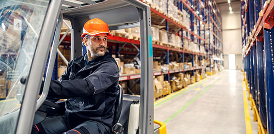 Ein Arbeiter mit Helm f&auml;hrt einen Gabelstapler durch ein gro&szlig;es Lager mit Regalen und Waren.