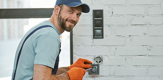 Ein Handwerker installiert eine Steckdose an einer Wand und blickt dabei zur Kamera.