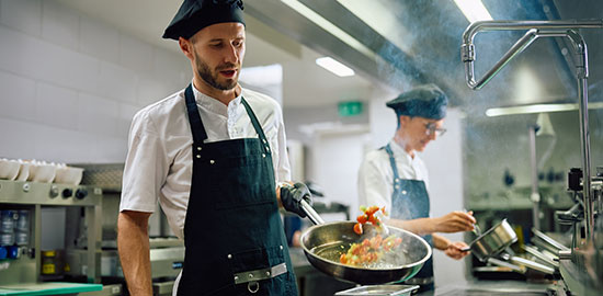 Ein Koch schwenkt eine Pfanne in einer professionellen K&uuml;che, w&auml;hrend im Hintergrund ein Kollege arbeitet.
