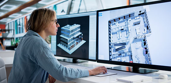 Eine Fachkraft arbeitet am Computer an digitalen Baupl&auml;nen und 3D-Modellen eines Geb&auml;udes.