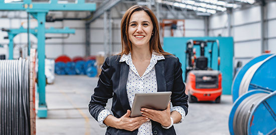 Eine Mitarbeiterin steht mit Tablet in einer Industriehalle und &uuml;berwacht Abl&auml;ufe.