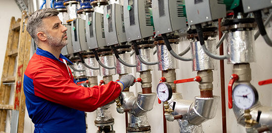 Ein Techniker &uuml;berpr&uuml;ft und wartet mehrere Heizungs- und Rohrsysteme in einem Technikraum.