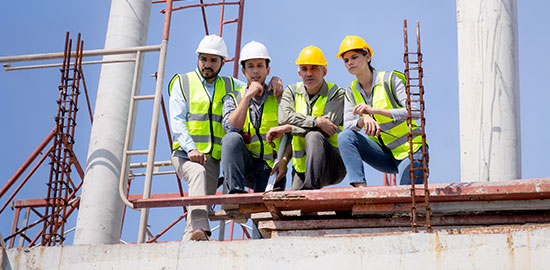 Mehrere Fachkr&auml;fte mit Helmen und Warnwesten sitzen auf einer Baustelle und besprechen Arbeitsschritte.