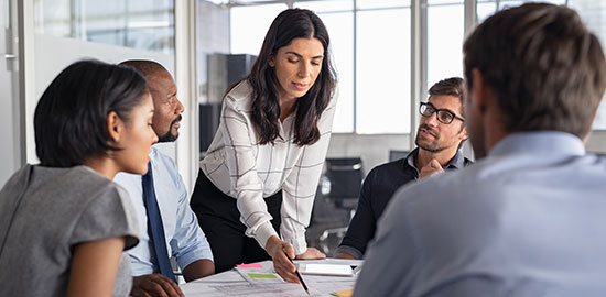 Eine Mitarbeiterin erkl&auml;rt Unterlagen in einem Meeting, w&auml;hrend ein Team aufmerksam zuh&ouml;rt.