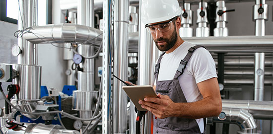 Ein Handwerker kontrolliert Rohrleitungen und technische Anlagen mit einem Tablet.
