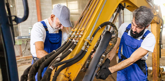 Zwei Mitarbeiter warten eine gro&szlig;e Baumaschine und pr&uuml;fen deren Bauteile.