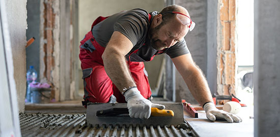 Ein Handwerker verlegt Fliesen und tr&auml;gt M&ouml;rtel auf einen Boden auf.
