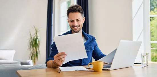 Ein Mann arbeitet am Laptop und pr&uuml;ft Unterlagen an einem Schreibtisch zu Hause.